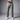 A man shown from the waist down wearing crater grey chinos with a brown leather belt, a black holstered handgun at the waist, and white sneakers, against a gray studio background.