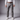 A man wearing crater grey chinos, a brown leather belt, a white long-sleeve button-up shirt tucked in, and white sneakers, shown from the waist down against a gray studio background.
