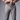 A close-up of a man's hands adjusting a black belt inside the waistband of crater grey chinos, beneath a brown leather belt, against a gray studio background.