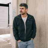 Man wearing a black jacket and light blue jeans standing against a textured wall.